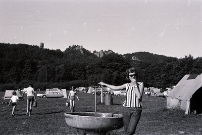 A young woman stands next to a public fountain in a campground setting. A large hillside and castle ruins are visible in the ...