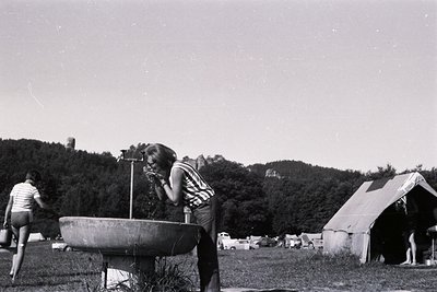 A young person leans over a drinking fountain on a grassy field. Background shows a tent, vehicles, and a forested hillside w...