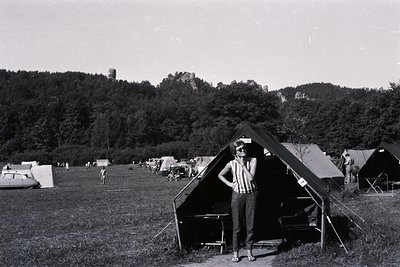 Young woman stands outdoors in front of a canvas tent at a campground. Several other tents and campers populate the grassy fi...
