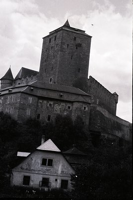 A dramatic black and white image showcasing a medieval castle complex atop a rocky escarpment. A tall, central tower dominate...