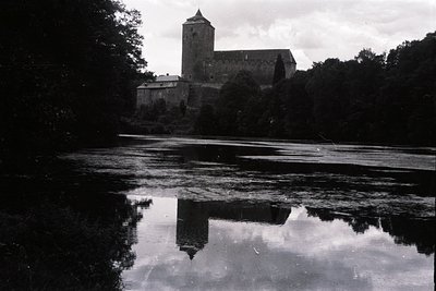 A stately, medieval castle rises above a tranquil lake, its reflection perfectly mirrored in the still water. Stone architect...