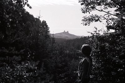 A solitary figure stands silhouetted within a forest, gazing at a distinctive rock formation dominating the distant landscape...