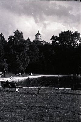 A view of a park, featuring a calm pond and tiered landscaping ascending to a structure on a hill. Benches line the water's e...