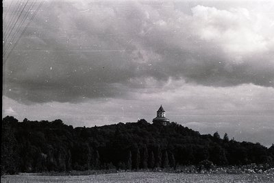 A stately building tops a densely wooded hill under a dramatic, cloudy sky. The structure appears to be a cupola or observato...