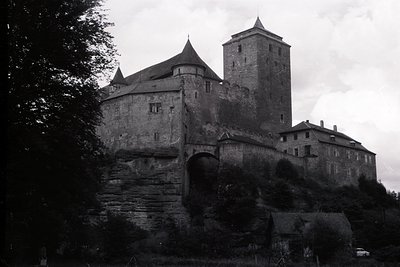 A dramatic, black and white view of a medieval castle perched atop a rocky outcrop. Features include a central tower, arched ...