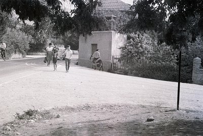 A family walks along a road alongside a small, stone building with a balcony. A man rides a donkey nearby. Likely rural scene...