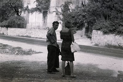 A man and woman converse on a rural road. The man, in work clothes, gestures while the woman carries a basket and water jug. ...