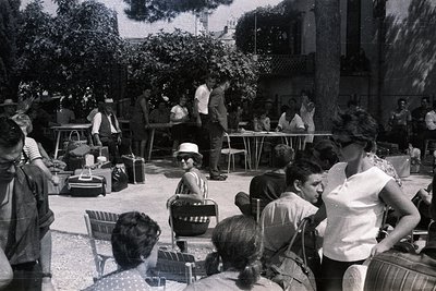 Group gathered outdoors, appearing to be awaiting transport. Suitcases and bags are prominent, suggesting travel. Architectur...