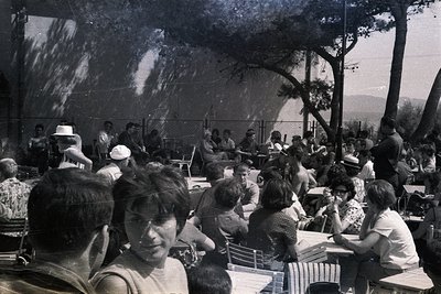 Black and white photograph captures a crowded outdoor cafe scene. Guests seated at tables, many with hats, enjoy the terrace ...