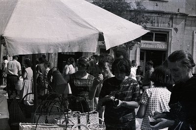B&W street scene, likely a market or fair. Vendors display woven baskets beneath a canvas awning. Shoppers, mostly women & ch...