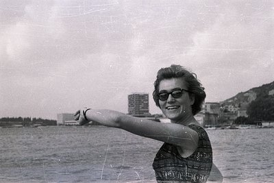 Young woman in patterned dress & cat-eye glasses extends arm towards the sea. Background features modern architecture and a h...