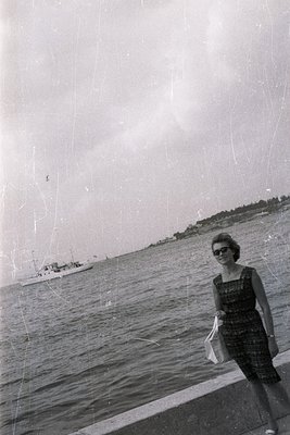 A woman in a dark dress and sunglasses stands near a seaside promenade, holding a fabric bag. A motorboat traverses the water...