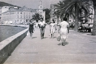 Four figures stroll along a seaside promenade, facing away from the camera. A formal building with a clock tower rises in the...