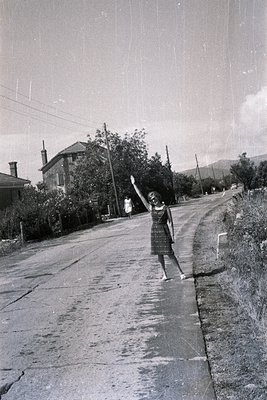 A young woman in a patterned dress waves while standing on a rural road. A stone house with a pitched roof and chimney is vis...