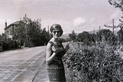 Black and white photo: a woman in a patterned dress & sunglasses stands roadside, beside a rural building, likely in the 1960...