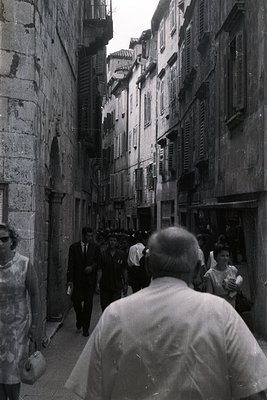 Narrow, cobbled street lined with tall, multi-story buildings featuring many windows. Pedestrians walk away from the camera, ...