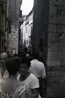 Crowded stone alleyway, likely a European coastal town. A group of people, mostly men and women, appear to be traversing the ...