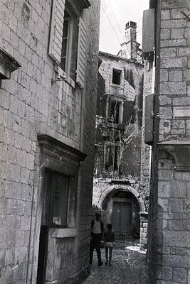 Narrow, cobbled alleyway flanked by stone buildings, likely Mediterranean. Two figures, a man and child, are walking away fro...