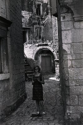 A young woman in a short dress and sandals poses within a narrow, stone-walled alleyway. Architectural details include an arc...