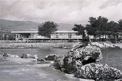 A young woman sits on a rocky breakwater, overlooking a seaside resort with a distinctive modernist architectural style. Pine...
