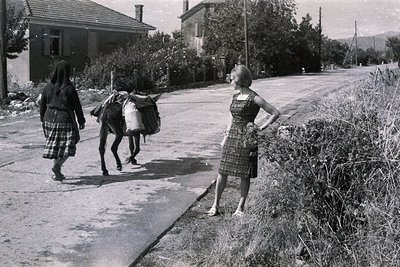 A woman in a traditional, dark dress and headscarf leads a donkey laden with goods along a rural road. A second woman in a pa...