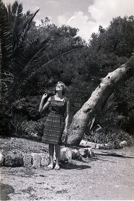 A young woman in a patterned, short-sleeved dress and sandals poses outdoors, leaning against a large, gnarled tree with visi...