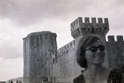A woman in 1960s attire, dark sunglasses, and a short bob haircut poses with a stone fortress. The structure features arched ...