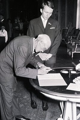 Elderly man in a suit signs a document at a table, assisted by a younger man in a suit and tie. Formal event, possibly a cere...