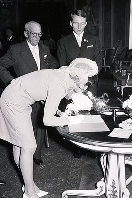 A woman in a veiled dress signs a guest book at a formal event. Two formally dressed men watch nearby. Likely a wedding or si...