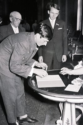 A man in a tailored suit signs a document on a decorative table, flanked by an older gentleman and another in a dark suit. Ap...