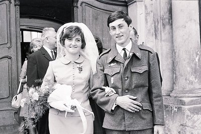 A smiling bride in a fitted dress and veil stands with a groom in military uniform. A bouquet is clutched in her gloved hand....