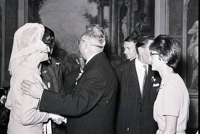 A formally attired man in a suit greets a woman in a veil, likely a bride, while two younger men and a woman wearing glasses ...