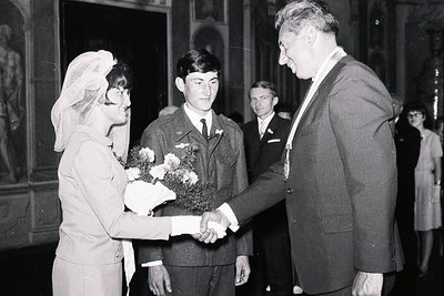 A bride in a headscarf receives a bouquet and shakes hands with a formally dressed gentleman who is wearing a military medal....