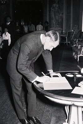 A formally dressed gentleman signs a document on a round table with a floral centerpiece. A marble bust and patterned carpet ...