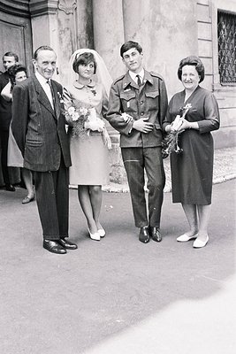 Formal portrait of a newly married couple and their families outside a building. Bride in a short dress, groom in military un...
