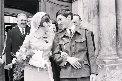 A newlywed bride in a short veil and gloves smiles warmly at a uniformed groom, likely a military officer. Architectural deta...