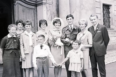 Black and white photo depicting a wedding party outside a stone building, likely 1960s. A bride in a veil holds hands with a ...