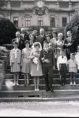 A formal portrait depicts a wedding party posed on stone steps before an ornate, multi-story building with a clock tower. Ten...