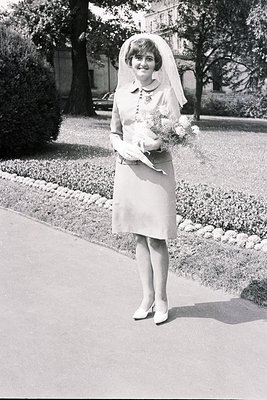 A young bride in a knee-length, A-line dress and veil poses outdoors. She holds a bouquet of flowers. Mid-century style; like...