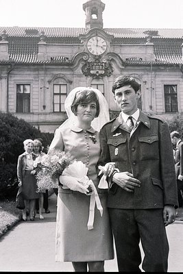 A newly married couple poses formally outdoors. The bride wears a short wedding dress with a veil and carries flowers. The gr...