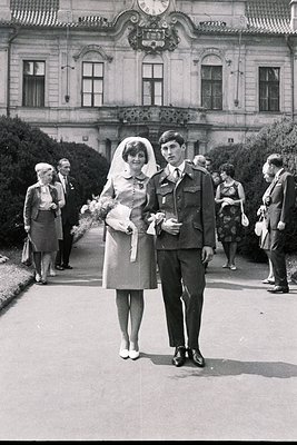 A newlywed couple poses formally outside a grand, neoclassical building. The bride wears a short, fitted wedding dress and ve...