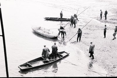 A group of men in military uniform are loading or unloading small boats from a sandy shoreline. Several men hold ropes, guidi...