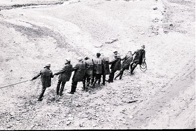 A group of men in military-style greatcoats and caps haul a rope across a rocky, uneven landscape. The scene suggests manual ...