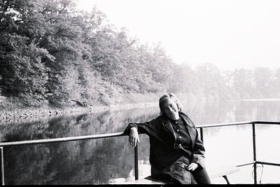 A young woman sits casually on a raised platform overlooking a body of water, likely a lake or wide river. Dense foliage line...