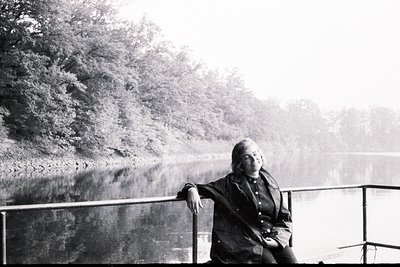 A seated woman in a dark, layered jacket overlooks a body of water framed by dense foliage. Likely a candid, mid-century port...