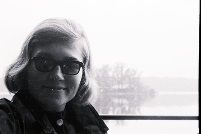 Close-up portrait of a woman with short, wavy hair and dark-rimmed glasses. She appears to be smiling slightly. A body of wat...