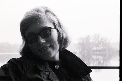 Portrait of a woman with short, styled hair and dark sunglasses. She wears a button-down collar and appears to be outdoors, p...