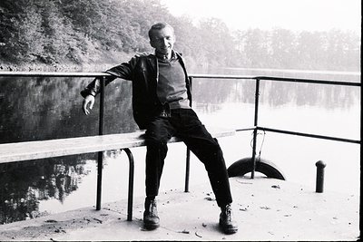 Man seated on a railing overlooking a tranquil lake with forested shoreline. He wears a dark jacket, sweater, and sturdy boot...