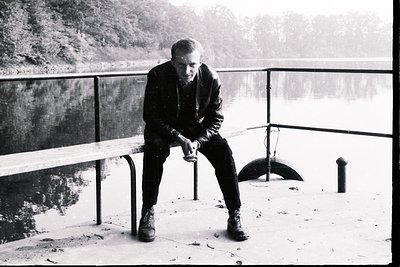Man seated on a concrete ledge overlooking water, dressed in a dark leather jacket and trousers, boots visible. Stark composi...