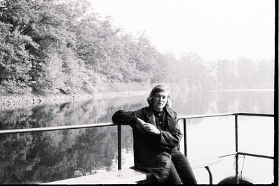Young man with long hair sits on a low railing overlooking a body of water, likely a lake or river, framed by dense foliage. ...